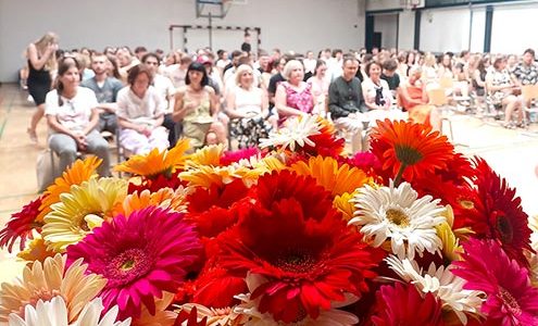 Podelitev maturitetnih spričeval - v ospredju šopek pisanih rož, v ozadju na stolih v telovadnici sedijo učitelji in maturanti.