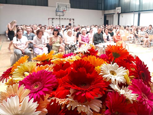 Podelitev maturitetnih spričeval - v ospredju šopek pisanih rož, v ozadju na stolih v telovadnici sedijo učitelji in maturanti.