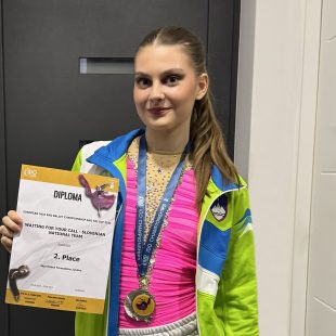 Tekmovalka stoji z medaljo okrog vratu in priznanjem v roki.