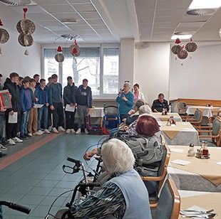 Dijaki nastopajo v domu starejših. Eden od dijakov igra na harmoniko, drugi pojejo.