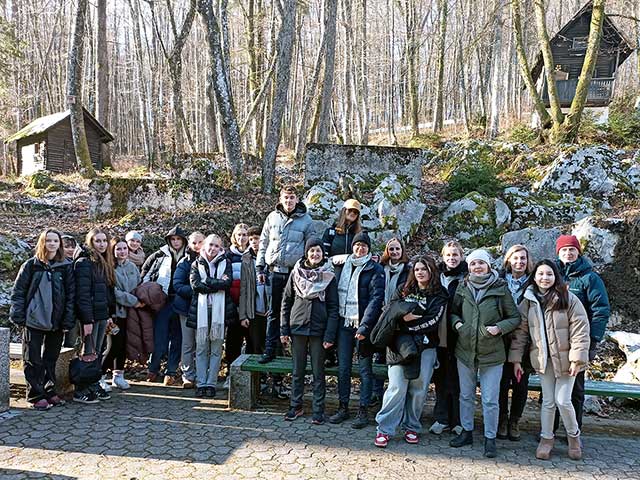 Skupinska fotografija udeležencev ekskurzije. Oblečeni so v tople jakne, kape in šale, stojijo pred pobočjem, ki ga prekriva gozd. Drevesa so zdaj brez listov, zato so vidna le debla in veje.